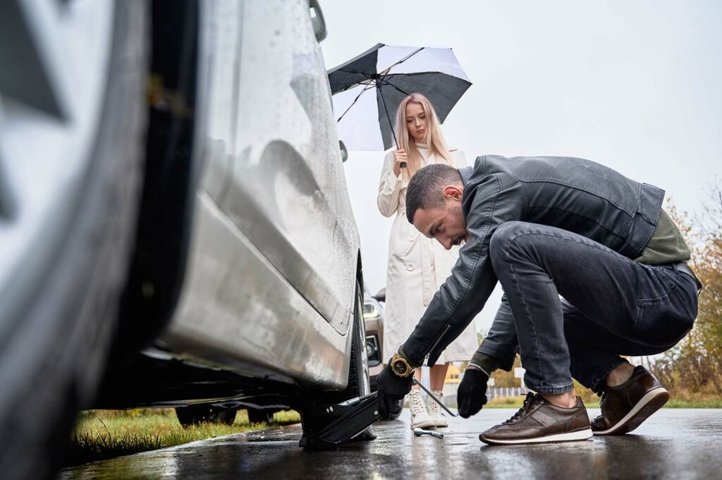 Charming blonde woman standing by automobile and holding umbrella while man placing car jack under vehicle. Handsome auto mechanic helping young woman to change flat tire on the road.